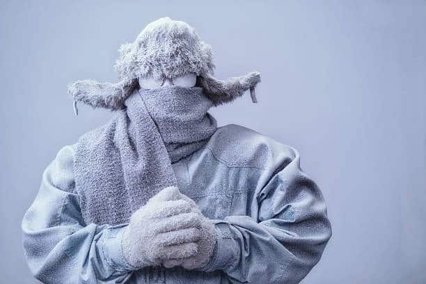 A frozen man with his hands clasped all bundled up in a fur trappers hat, scarf, parka and knit gloves, covered in snow and frost trying to stay warm on a very cold gray Winter's day.